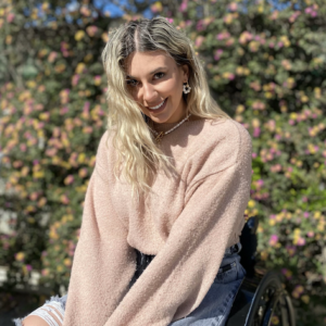 Portrait of Dimitra. She is wearing a pink sweater and jeans, smiling at the camera while seated in her wheelchair. Behind her is a wall covered with blooming flowers.