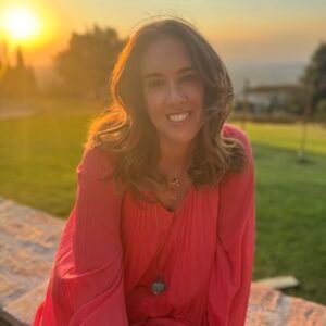 Portrait of Ileana. She is wearing a coral, flowy dress and smiling at the camera. Behind her is a countryside landscape at sunset