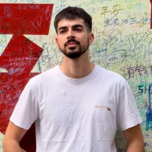 Portrait of Konstantinos. He is wearing a white T-shirt and has a natural, neutral expression. Behind him is a wall featuring phrases written in Japanese